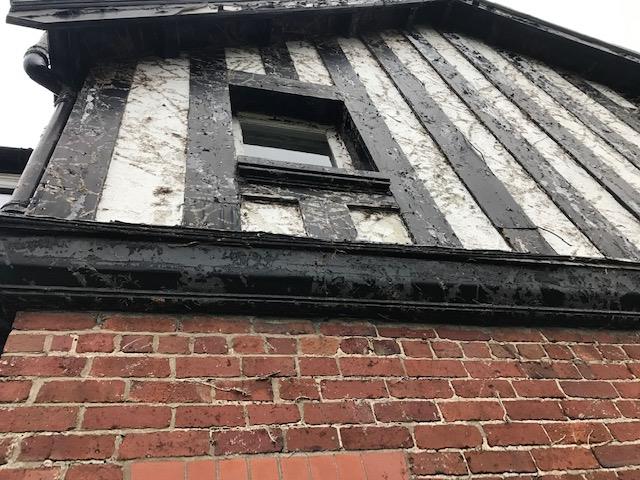 A close-up, low-angle photograph of the upper exterior of an existing house, showing red brickwork below and weathered black-and-white half-timbering above.