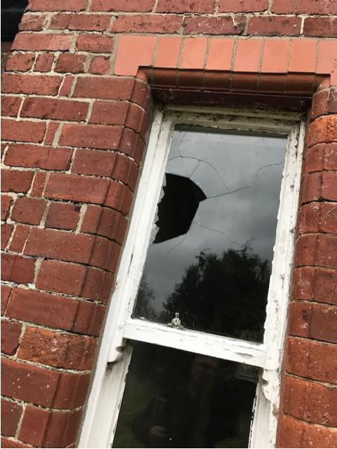 A close-up photograph of a red brick wall featuring a white-framed sash window with a large broken pane.