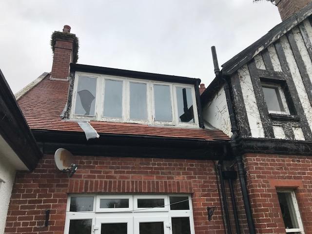 A low-angle photograph showing the upper facade of a brick house with a tiled roof and a dormer window. A white garage door is visible below, and a half-timbered neighbor stands to the right.