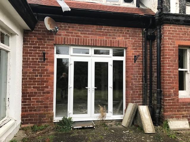 A photograph showing the exterior brick wall of a residential property featuring a large white uPVC window and door unit with construction materials on the ground.