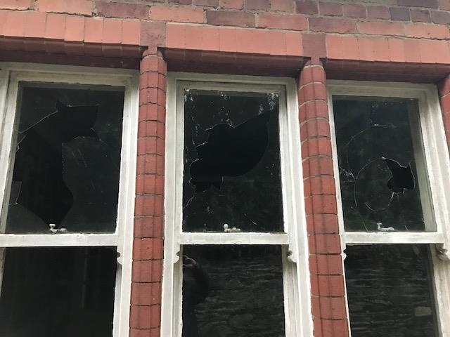 A close-up photograph of a red brick building facade featuring three sash windows with significant glass breakage and damage.