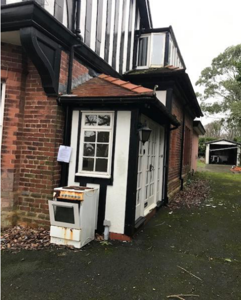 Exterior photograph of a residential property featuring a brick and half-timbered facade with a white porch extension and a garage visible in the background driveway.
