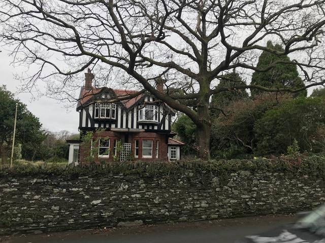 A photograph of an existing detached house featuring a black and white timber-framed upper story and brick lower story, situated behind a stone wall.