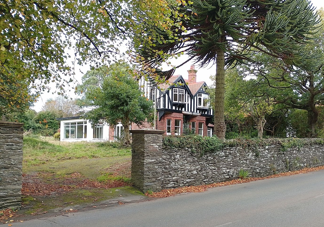 A street-level photograph showing a large detached house with black and white timber framing and a white extension, situated behind a stone wall and surrounded by mature trees.