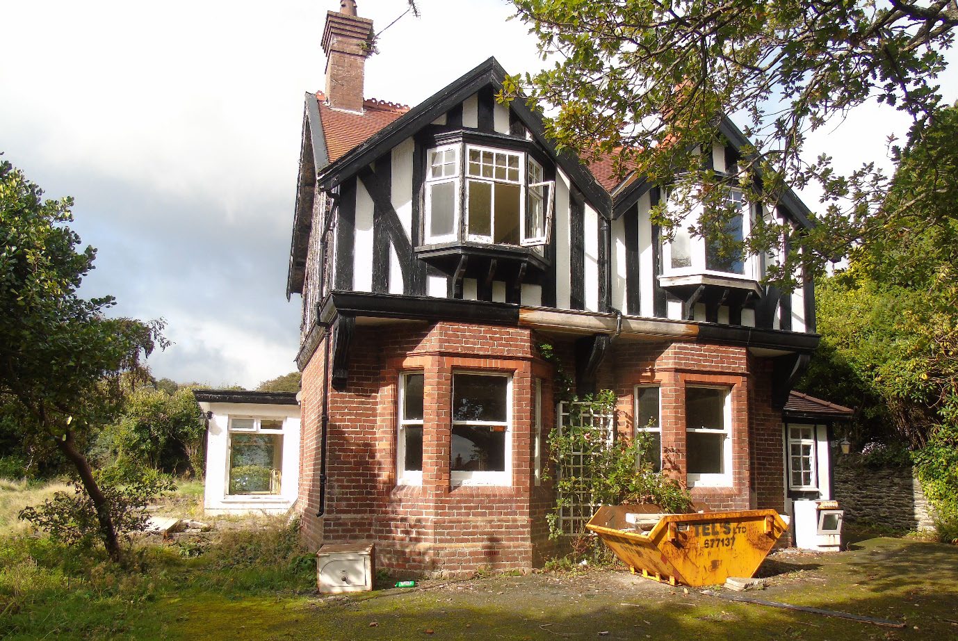 A photograph of a large, two-story detached house featuring red brick on the ground floor and black-and-white timber framing on the upper level. A yellow skip is visible in the front garden, suggesting preparation for...