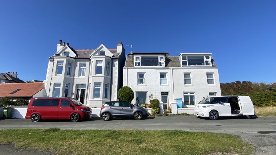 A street-level photograph showing a white detached house next to a building with large dormer windows, with several vehicles parked on the road.
