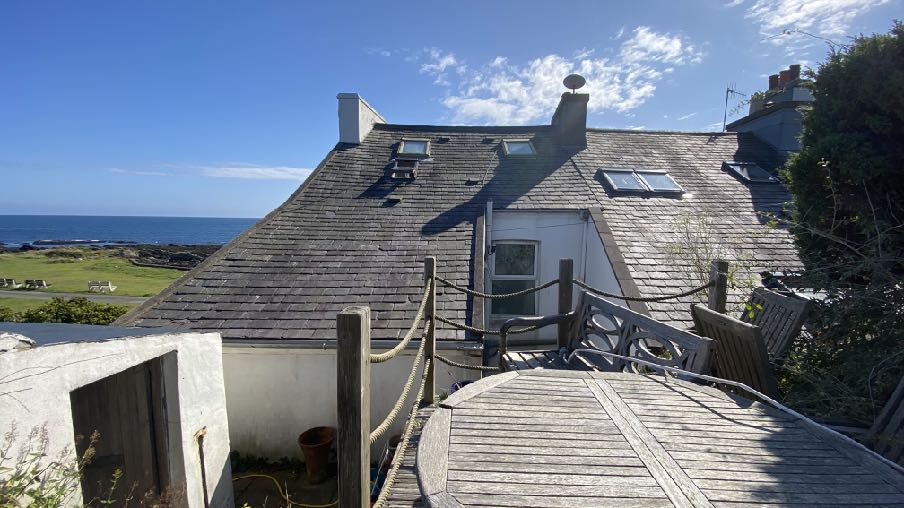 A photograph showing the slate roof of a house featuring skylights and dormer windows, viewed from a wooden deck with a coastal background.