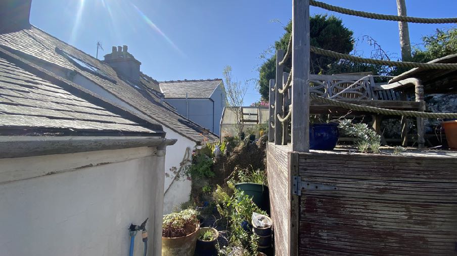 A photograph showing the side and rear of a white-walled house with a slate roof and skylights, next to a wooden deck and garden area.