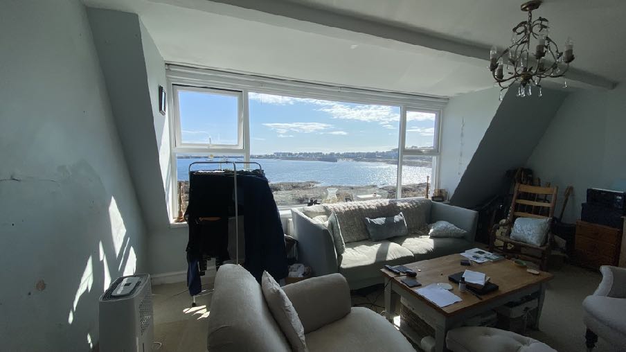 Interior photograph of a living space with large windows overlooking the sea and a sloping ceiling on the right.