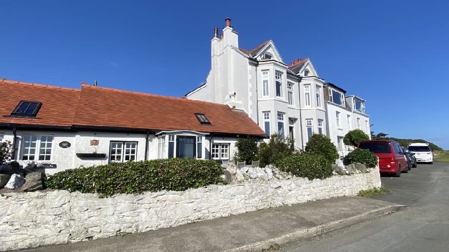 A street-level photograph showing a white single-story bungalow with a red tiled roof and dormer windows, situated next to a larger multi-story house.