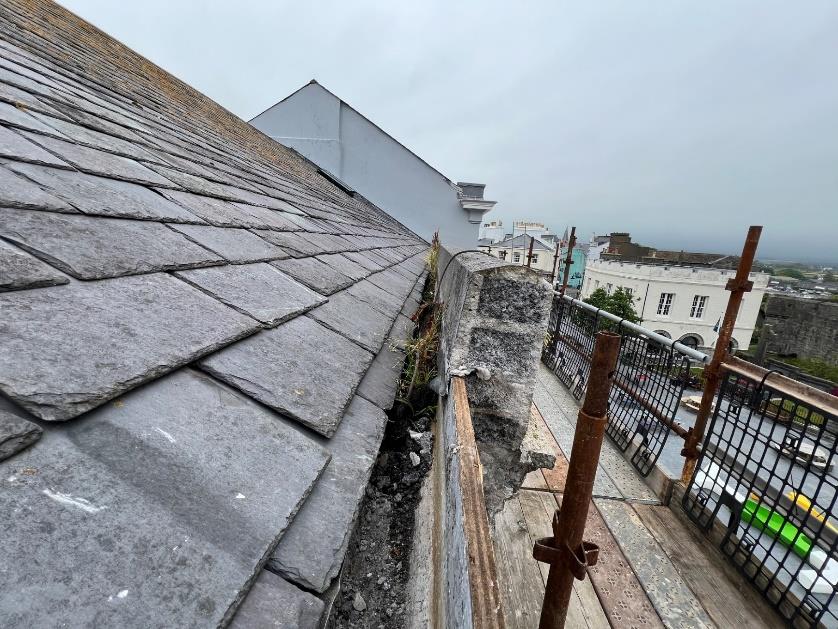 A high-angle photograph showing a slate roof edge with a stone parapet and gutter area, surrounded by scaffolding poles and wooden planks. The background shows a coastal town scene with other buildings and the sea.