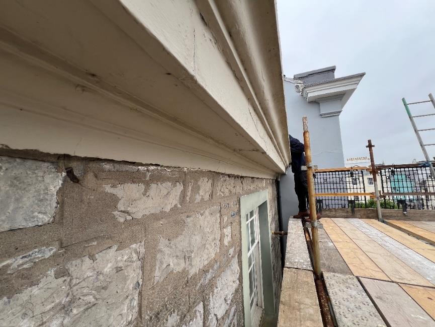 A close-up photograph showing the exterior stone wall and white cornice of a building under repair, with scaffolding planks visible along the side.