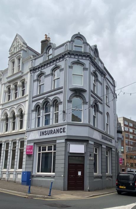 A street-level photograph of a grey, three-story corner building with 'INSURANCE' signage on the ground floor, situated in an urban area.