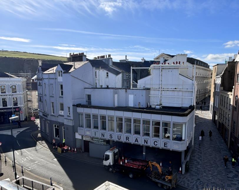 A high-angle photograph of a street corner featuring a white commercial building with 'INSURANCE' signage and construction vehicles parked in front.