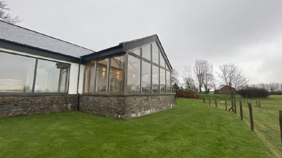 A photograph showing a detached house with a large, modern glass extension or conservatory featuring a stone plinth, set within a grassy rural garden.