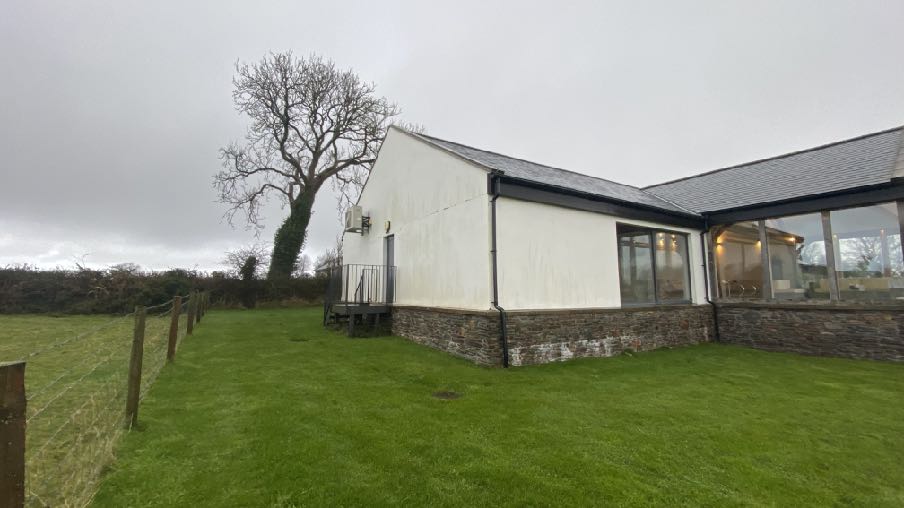 A photograph showing the side and rear elevation of a white single-story building with a stone plinth, featuring a wooden deck and a large grassy lawn in a rural setting.