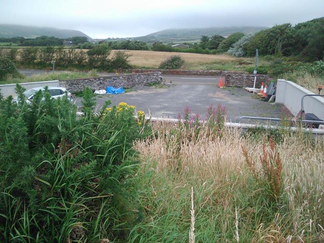 A photograph of a rural site featuring a paved driveway or parking area enclosed by stone walls, with overgrown vegetation in the foreground and hills in the distance.