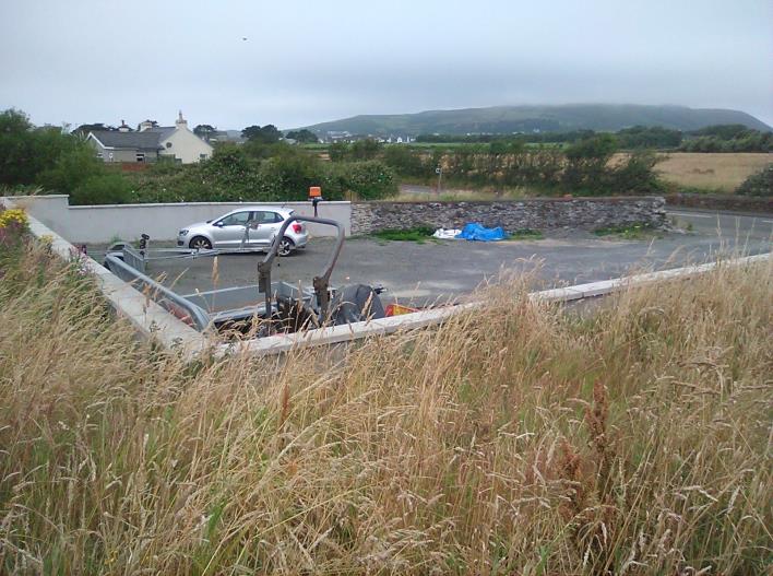 A photograph showing a paved driveway area with a silver car, bordered by tall grass in the foreground and rural hills in the background.