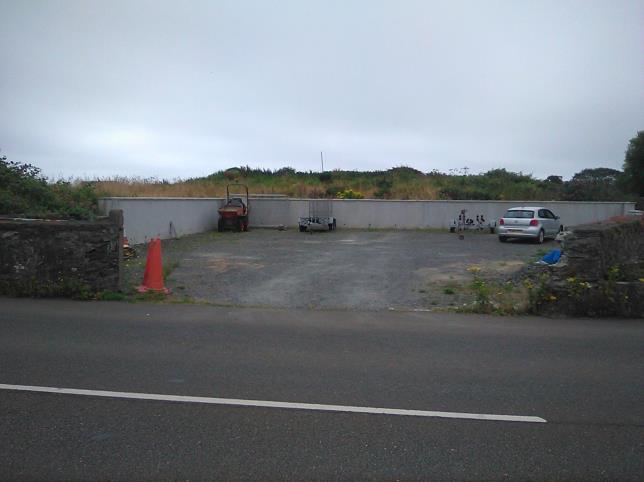 A photograph showing a paved driveway and parking area bordered by stone and concrete walls, viewed from the roadside.