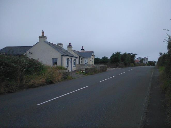 A street-level photograph showing existing white-walled houses with slate roofs alongside a paved road in a rural setting.
