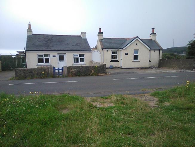 A street-level photograph showing two white, single-story bungalows situated behind a low stone wall and road verge.