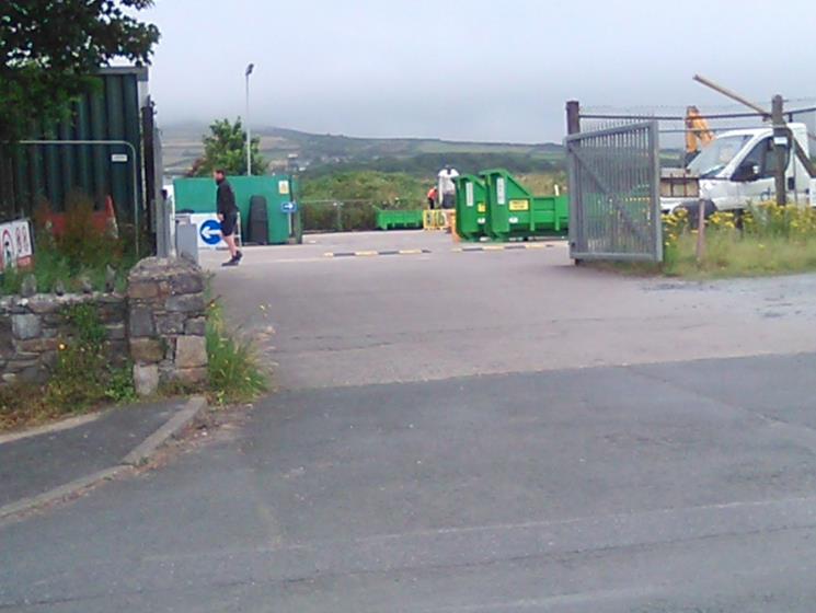 A photograph of a paved yard or entrance featuring large green waste skips, a white truck, and a metal gate, with a rural landscape in the distance.