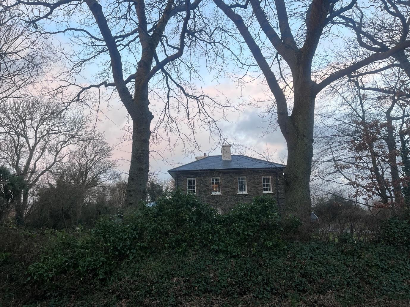 A photograph showing a stone building with a slate roof and white sash windows, partially obscured by large bare trees and dense vegetation. The setting appears to be rural, consistent with the Andreas parish location.