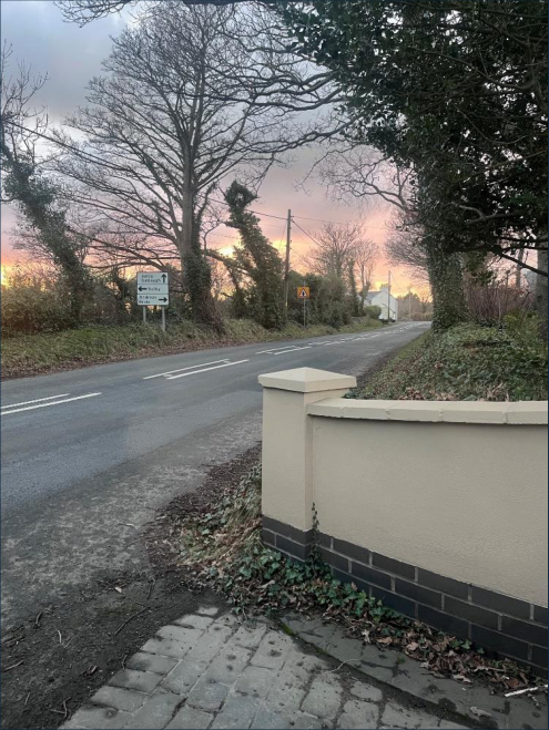 A photograph showing a roadside view with a low cream-colored boundary wall in the foreground and a road stretching into the distance under a sunset sky.