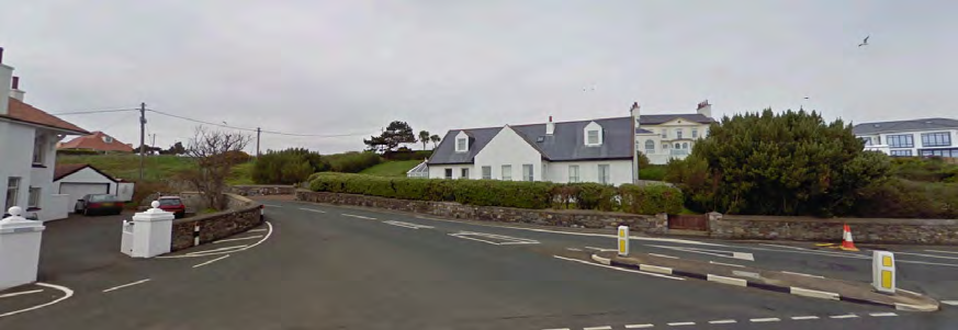 A panoramic photograph showing a road junction with traffic calming measures and residential properties in the background, including a white house behind stone walls and hedges.