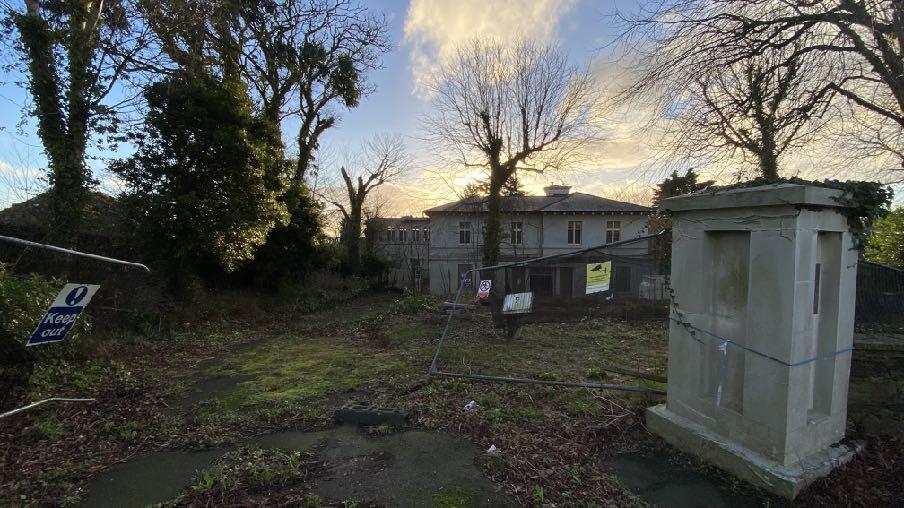 A photograph showing a grassy rear garden area with a fence, a concrete utility structure, and a large two-story house in the background surrounded by trees.
