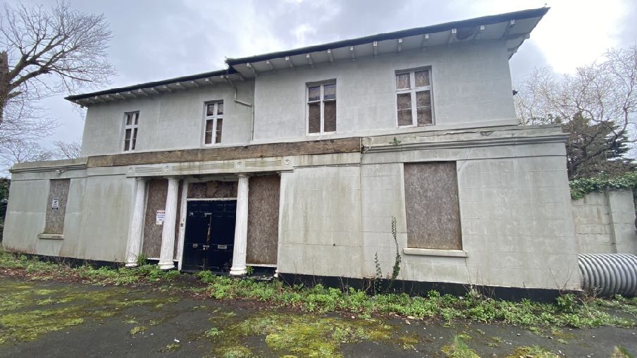 A photograph showing the front elevation of a two-story detached building with a lower single-story section featuring columns. The ground floor windows are boarded up, indicating disuse or ongoing renovation work.