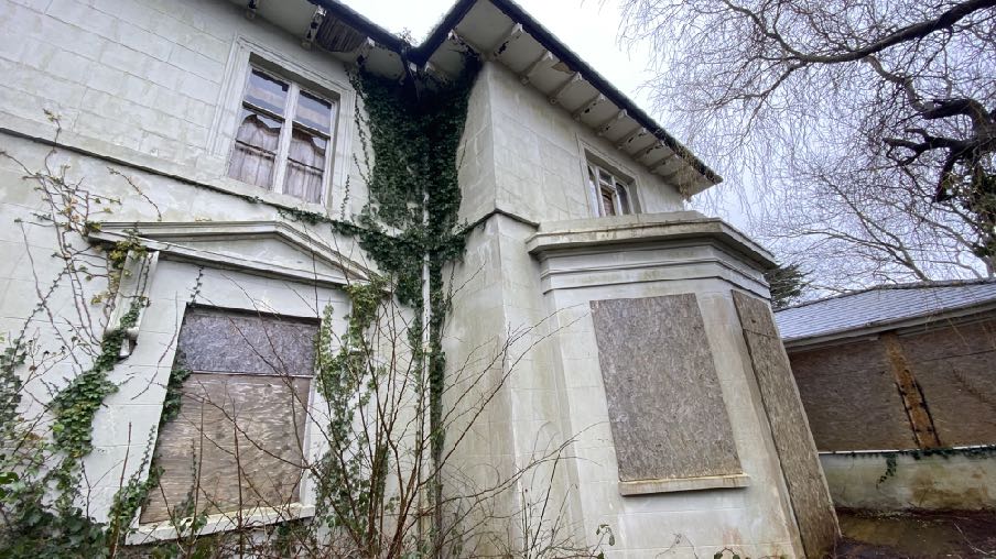 A low-angle exterior photograph of a white stone building featuring boarded-up windows and climbing ivy.