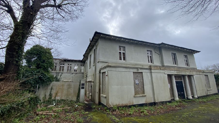 A photograph showing a large, dilapidated two-story building with boarded windows and overgrown vegetation, likely a former commercial or public building.