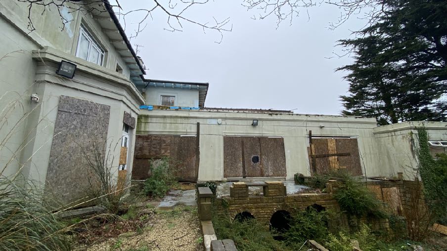 A photograph showing the rear elevation of a property featuring a flat-roofed extension with boarded windows and an overgrown garden area.