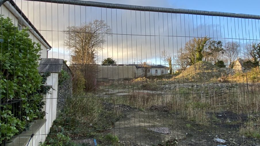 A photograph taken through a wire fence showing an overgrown rear plot with a pile of rubble and a white house in the background.