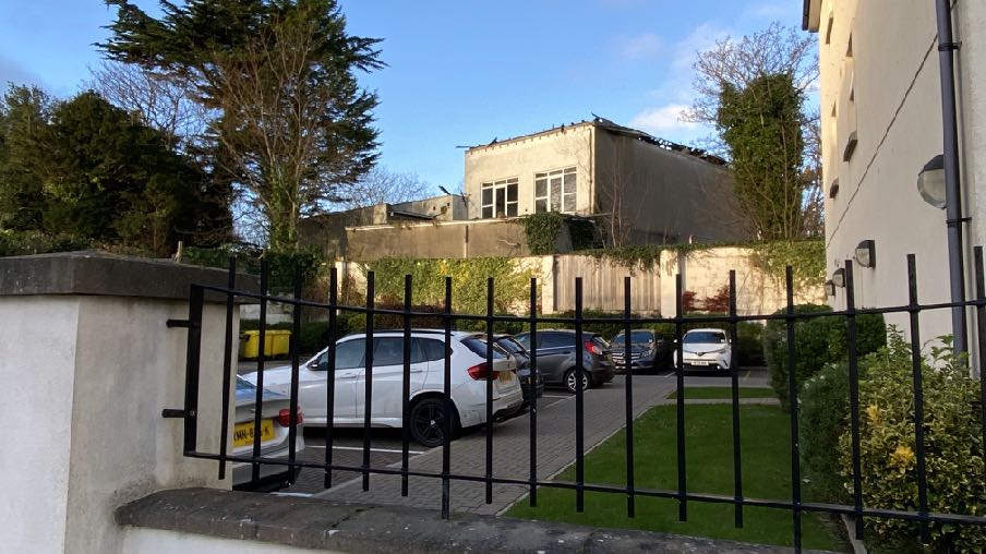 A street-level photograph showing a parking area with cars behind a black metal fence, with a two-story building structure visible in the background.