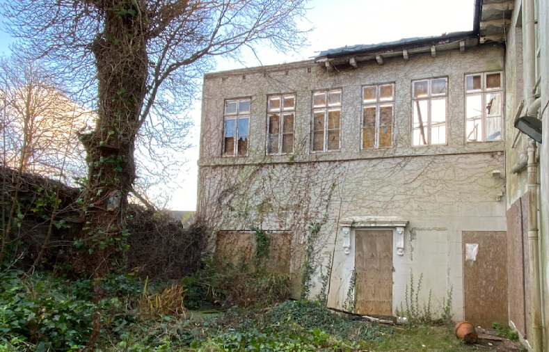 A photograph showing a dilapidated two-story rear extension with boarded windows and ivy-covered walls.