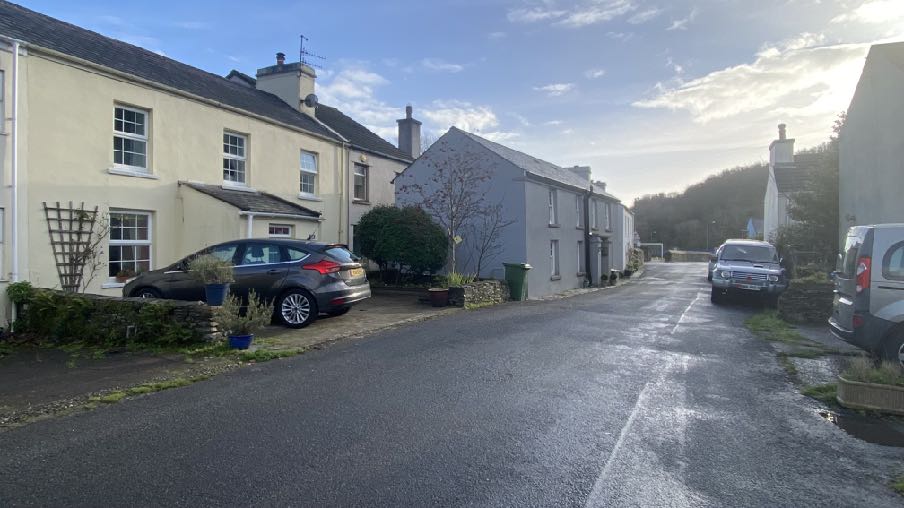 A street-level photograph showing a row of residential houses in a rural village setting with parked cars and a hill in the background.