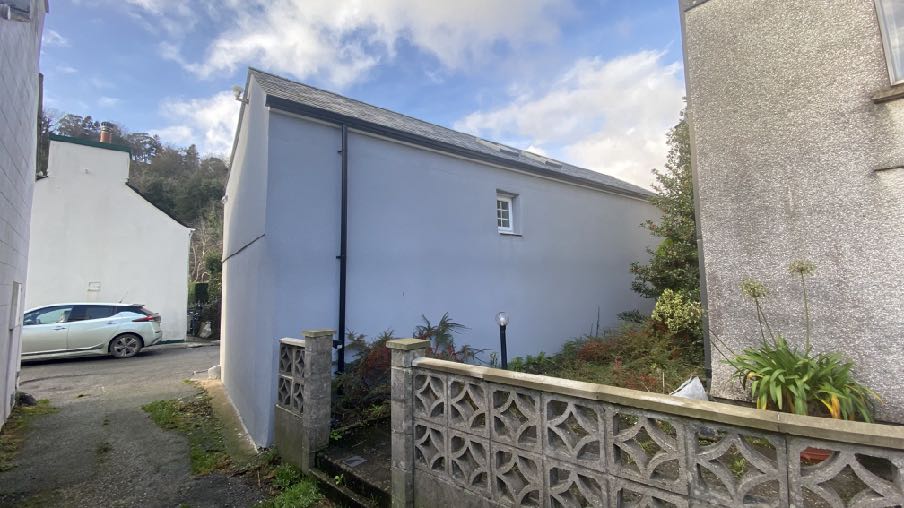 A photograph showing the side elevation of a white residential building with a slate roof, situated next to a parked car and a decorative concrete fence.