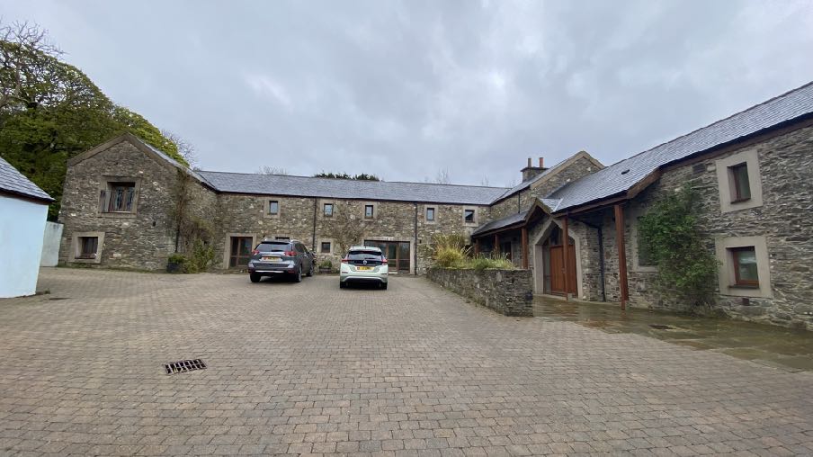 A photograph showing a large stone building complex with a paved driveway and two parked cars, set in a rural environment.