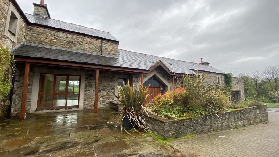 A photograph showing the exterior of a stone-built detached dwelling with a modern glass extension or patio area on the left side. The property features a paved patio, a low stone wall with landscaping, and a driveway...