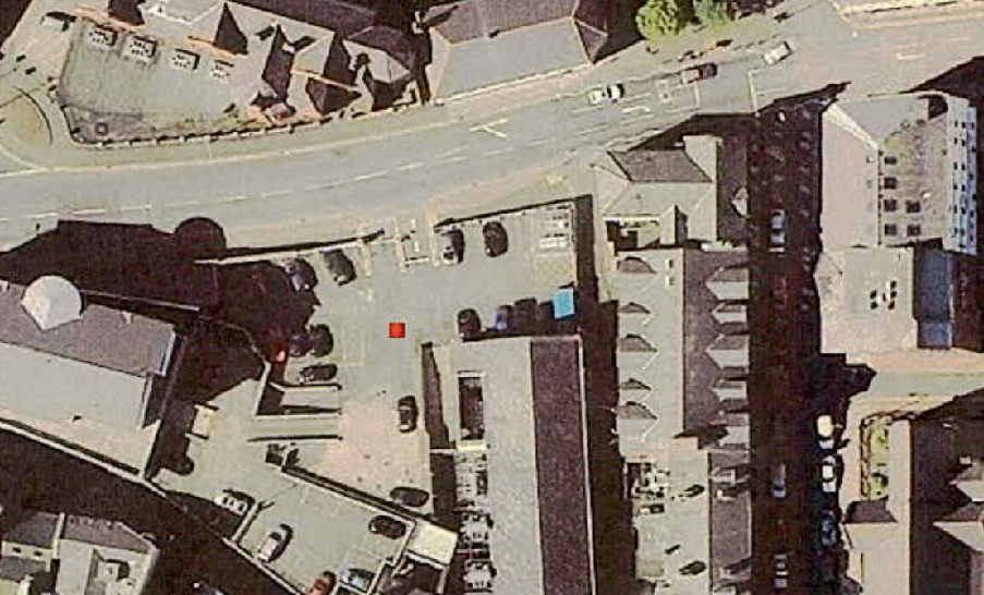 An aerial site plan showing a paved parking area with a red marker indicating the application site, situated alongside a road and adjacent buildings.