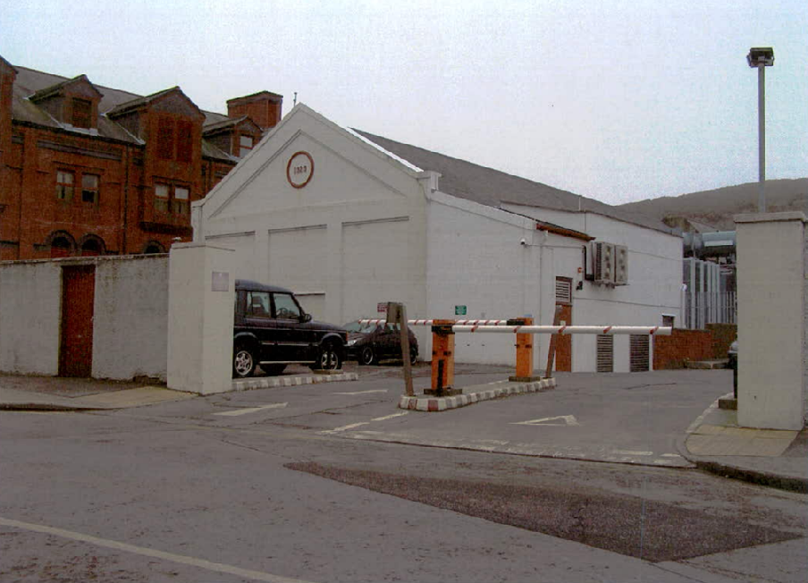 A photograph showing the entrance to a facility with a white gabled building, a barrier arm, and parked vehicles.