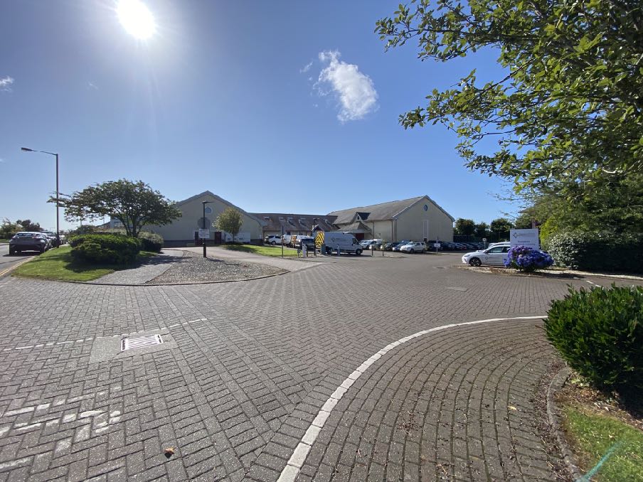 A sunny exterior photograph showing a paved parking area and driveway leading to a large, light-colored commercial or institutional building complex.