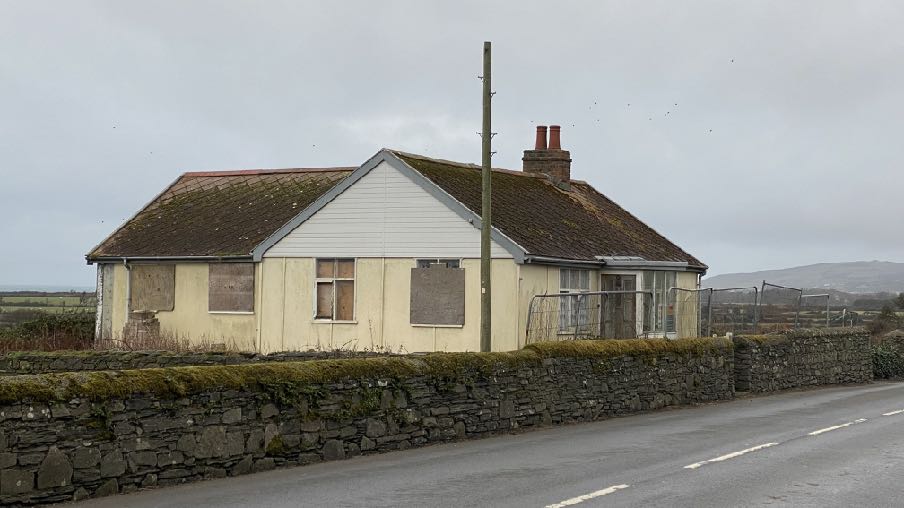 A photograph of a single-story cream bungalow with boarded windows and a conservatory extension, situated behind a stone wall along a rural road.