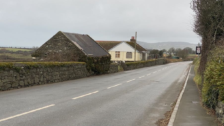 A street-level photograph showing a rural road with a stone wall, a dilapidated stone outbuilding, and a white bungalow in the background.