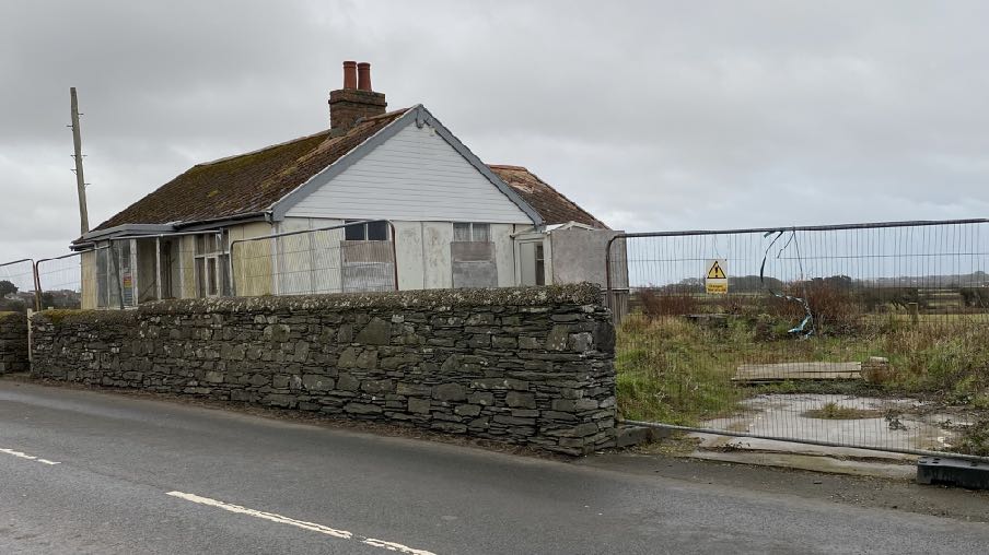 A street-level photograph showing a single-story detached bungalow situated behind a stone wall and wire fence, likely the existing property to be replaced.