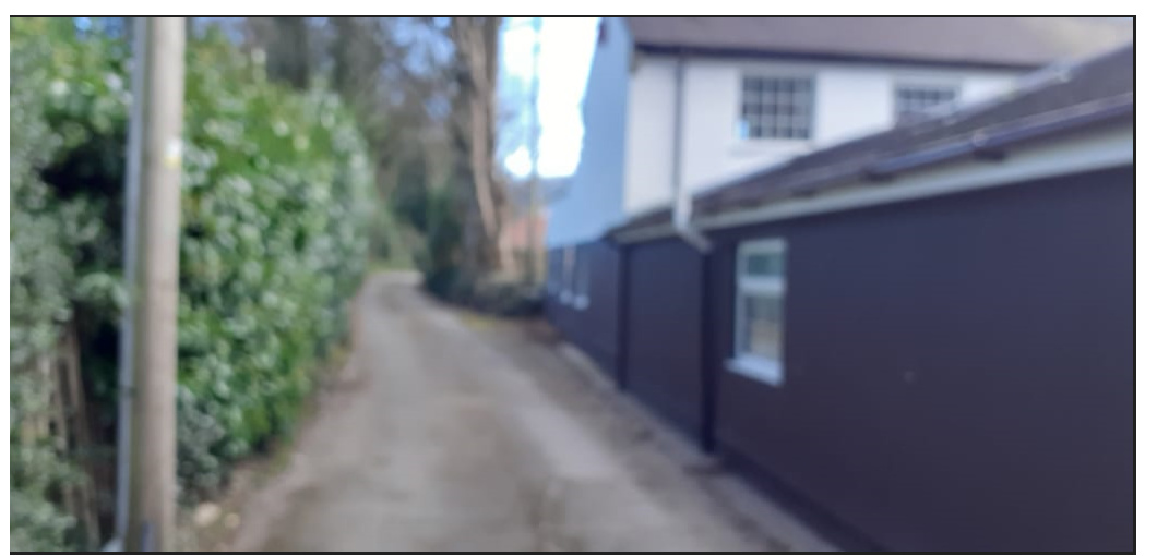 A slightly blurry photograph showing a narrow lane or driveway bordered by a tall hedge on the left and a white house with a dark extension on the right.