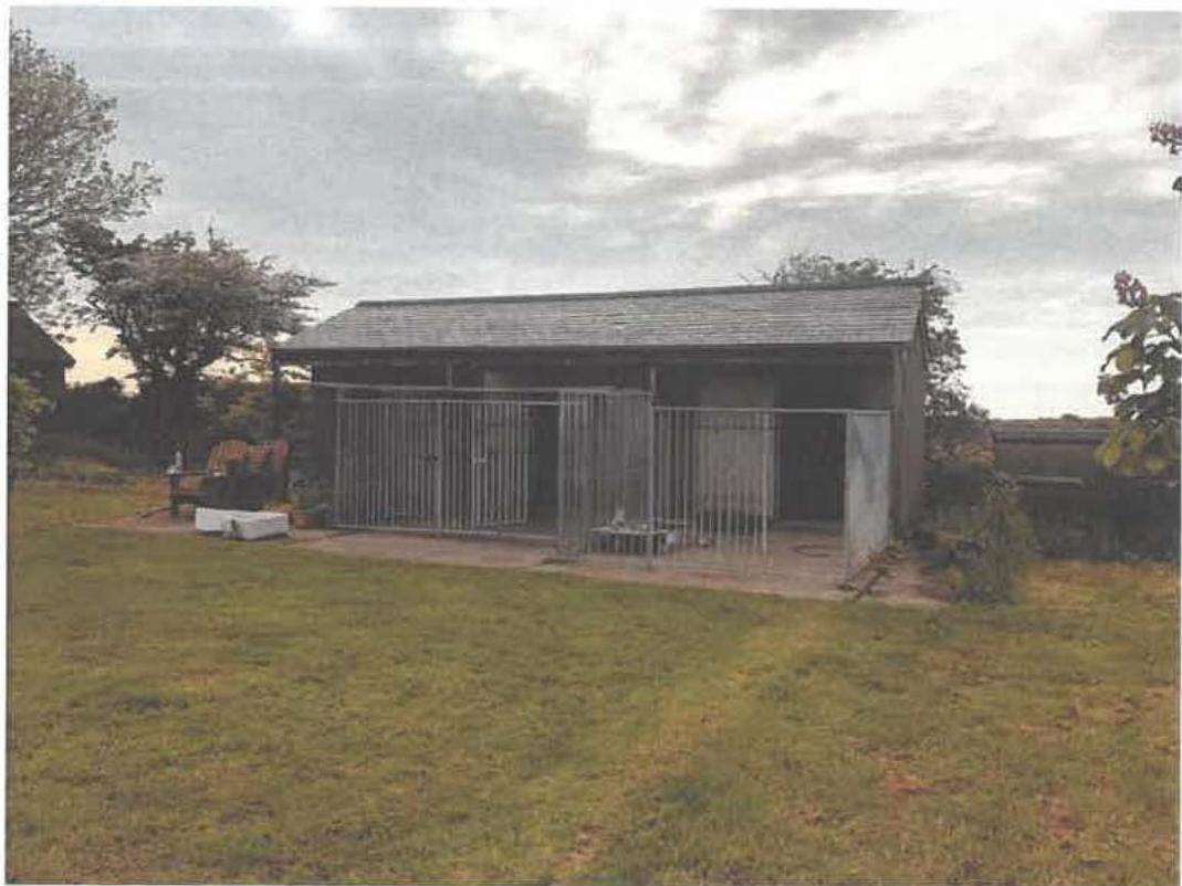 A photograph showing a long, single-story stable block with metal railings along the front, situated in a grassy rural setting.