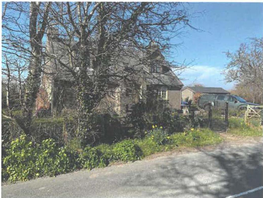 A photograph showing a stone cottage or house partially obscured by trees in a rural setting, with a road in the foreground and outbuildings visible in the background.